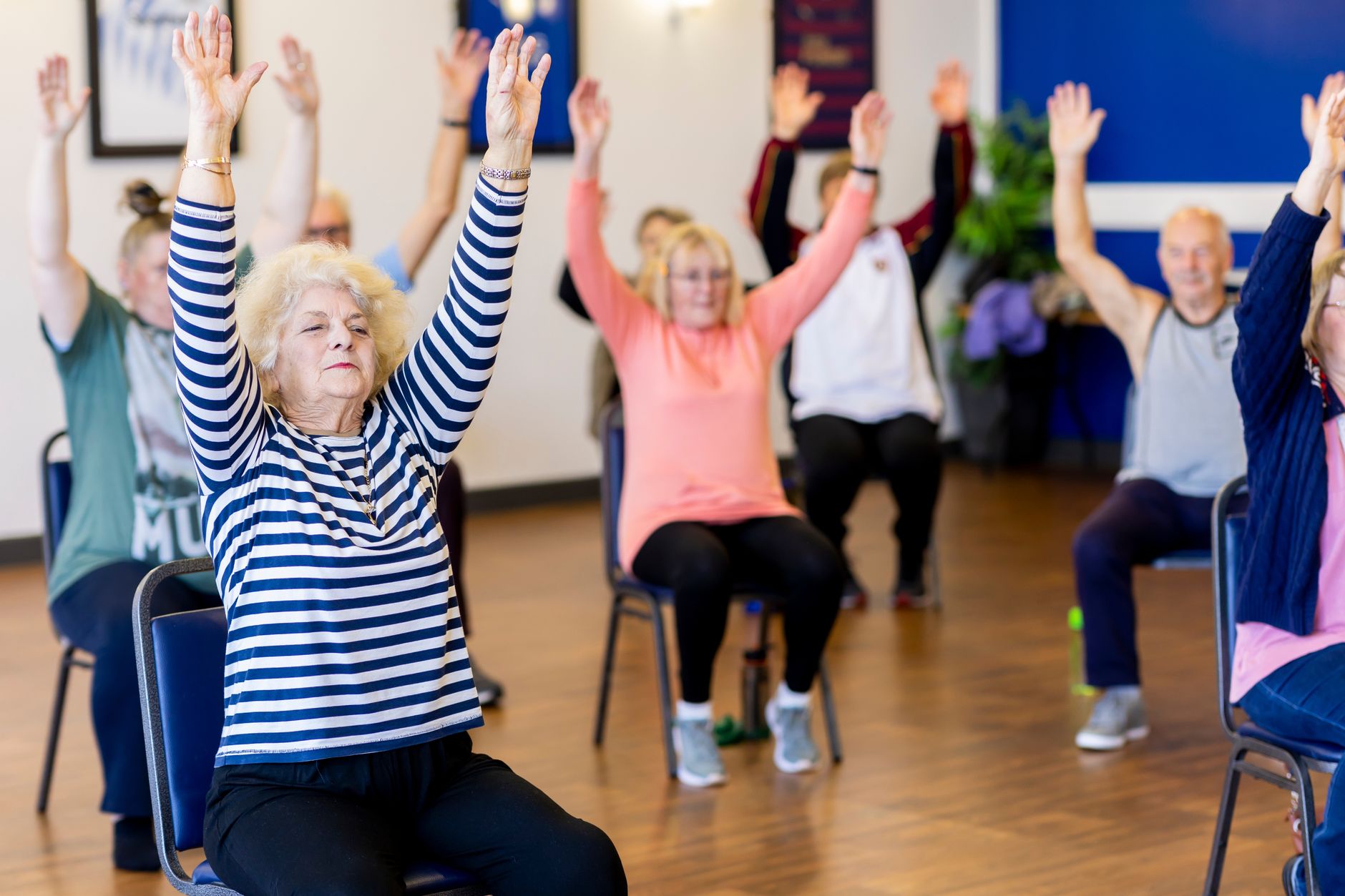 A group of older adults seated on chairs raise their arms together during an exercise class in a bright room