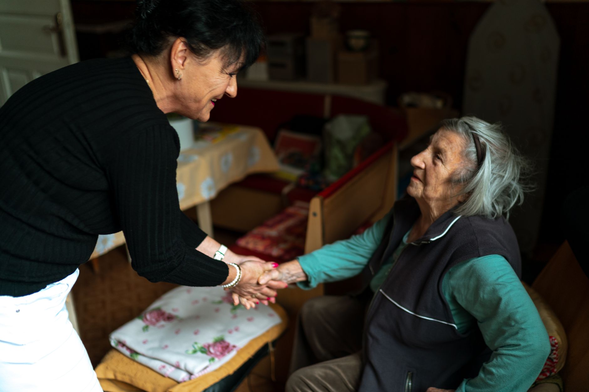 A smiling woman leans in to take the hand of an older woman seated in an armchair at home