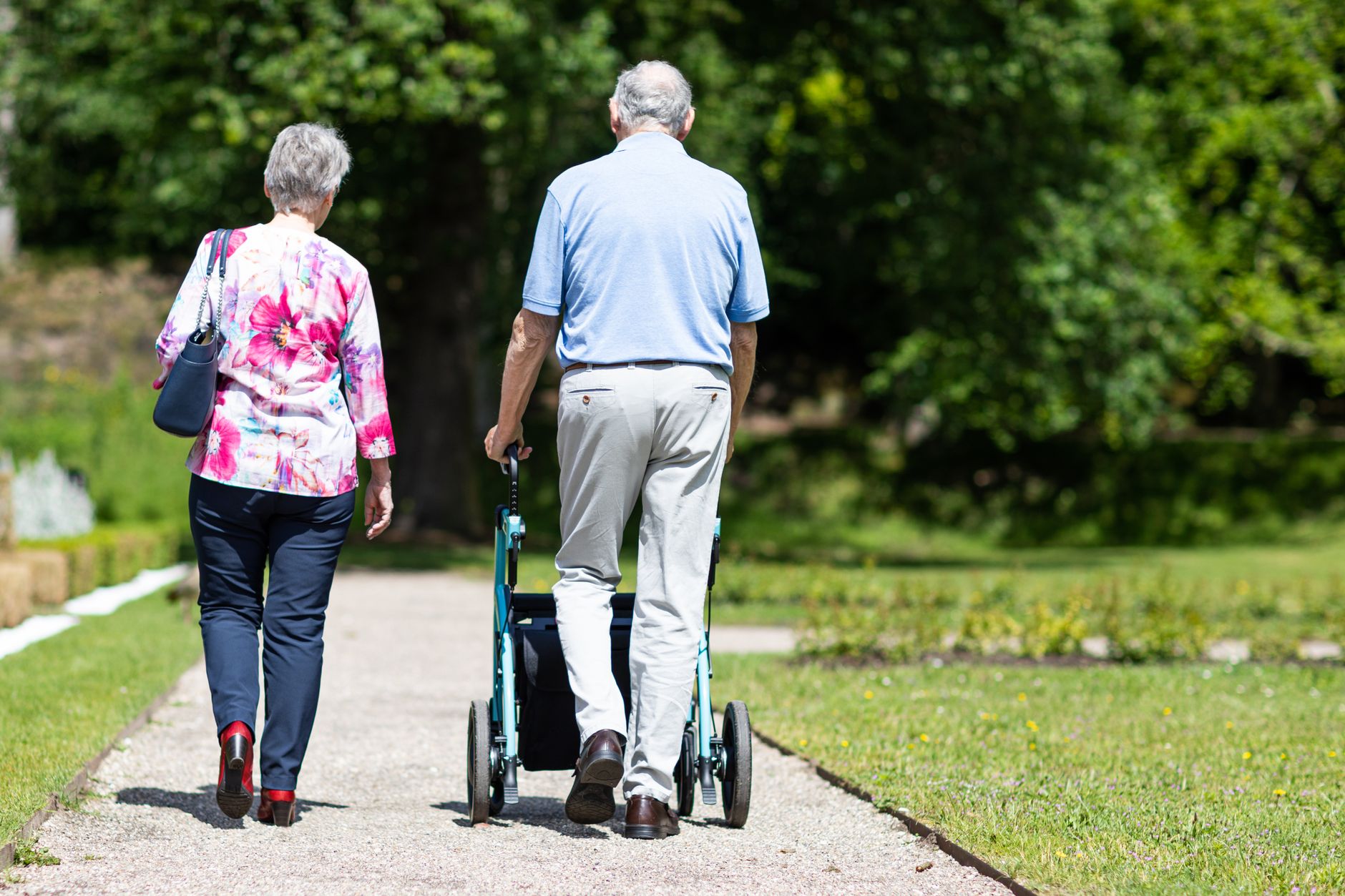An older woman in a floral blouse and an older man pushing a light blue rollator walk side by side down a gravel garden path lined with grass and trees