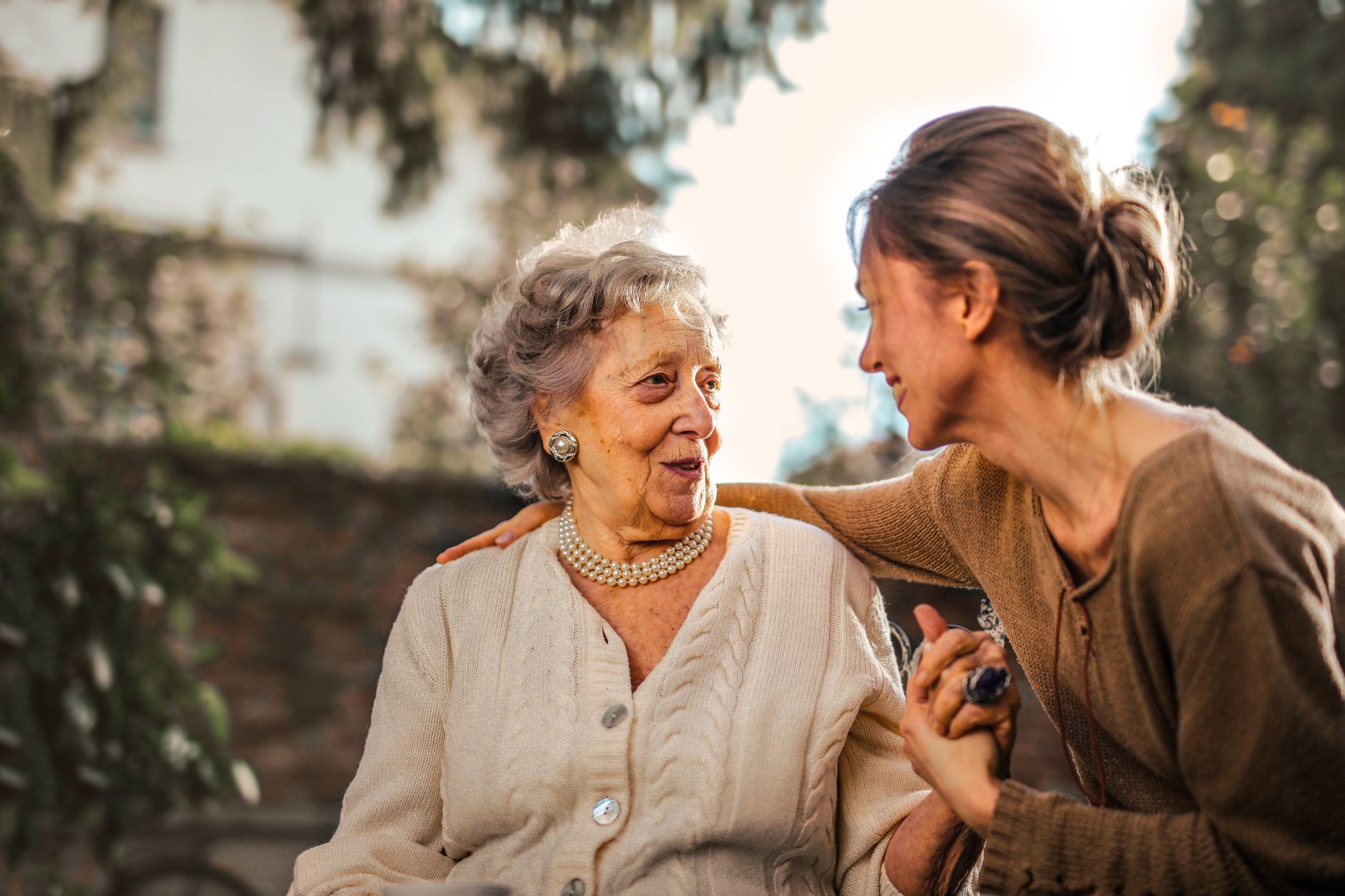 A smiling woman leans in with her arm around an older woman's shoulders as they hold hands outdoors