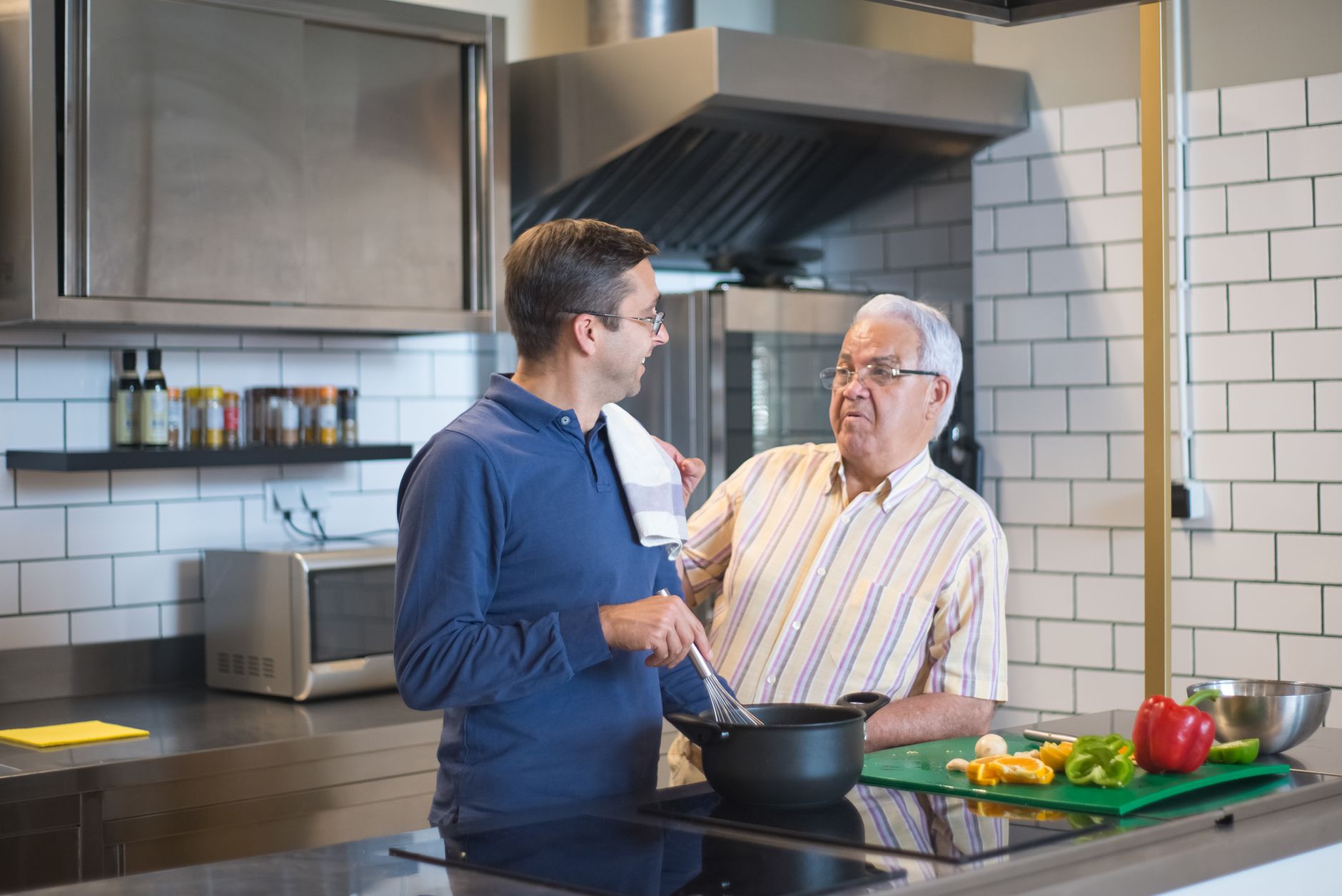 An older man in a striped shirt rests his hand on the shoulder of his adult son, who stirs a pot at a stainless steel kitchen counter with fresh peppers on a cutting board