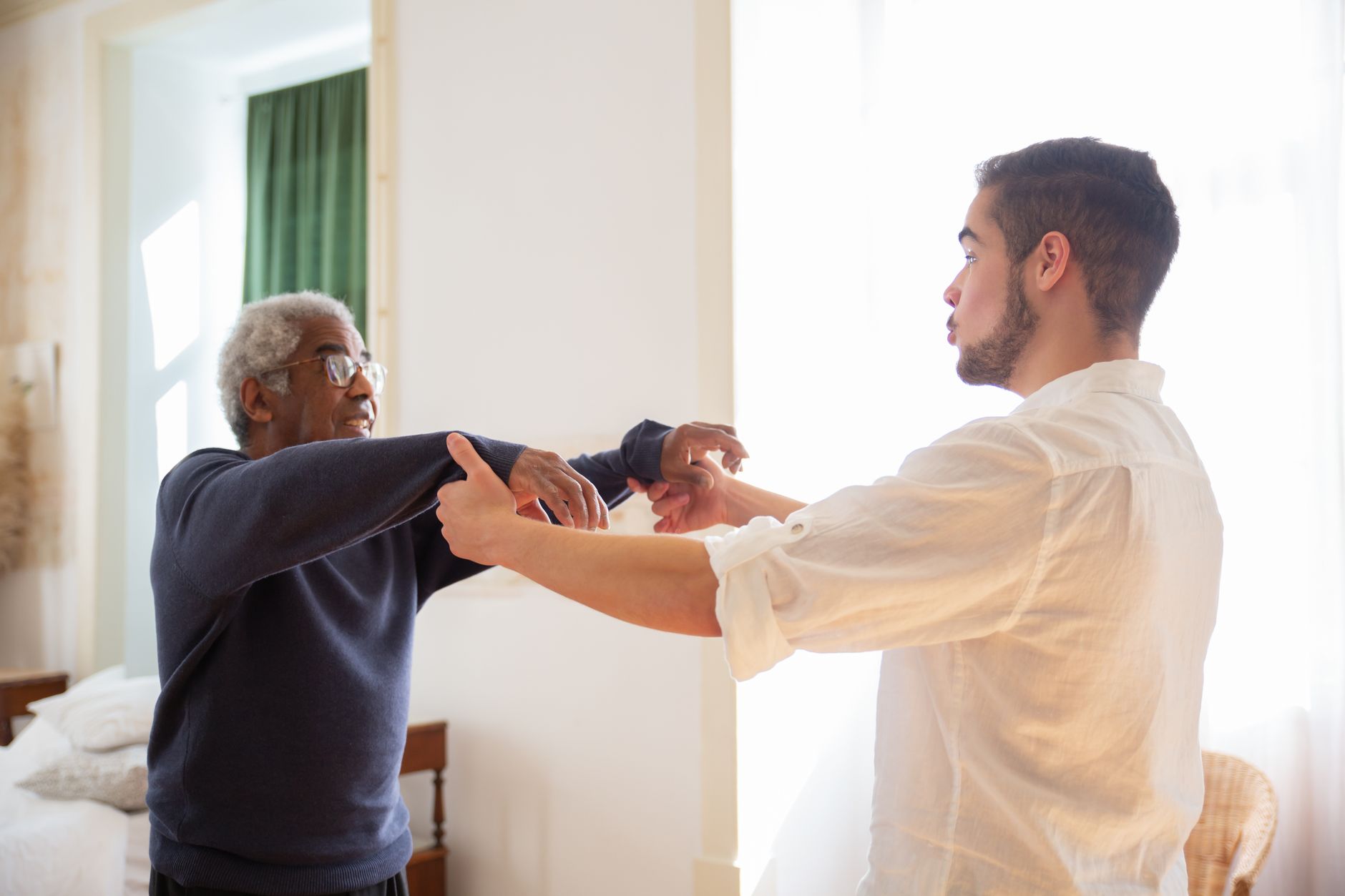 A smiling older man in glasses and a navy sweater stretches his arms forward with help from a younger man in a white shirt in a bright, sunlit bedroom