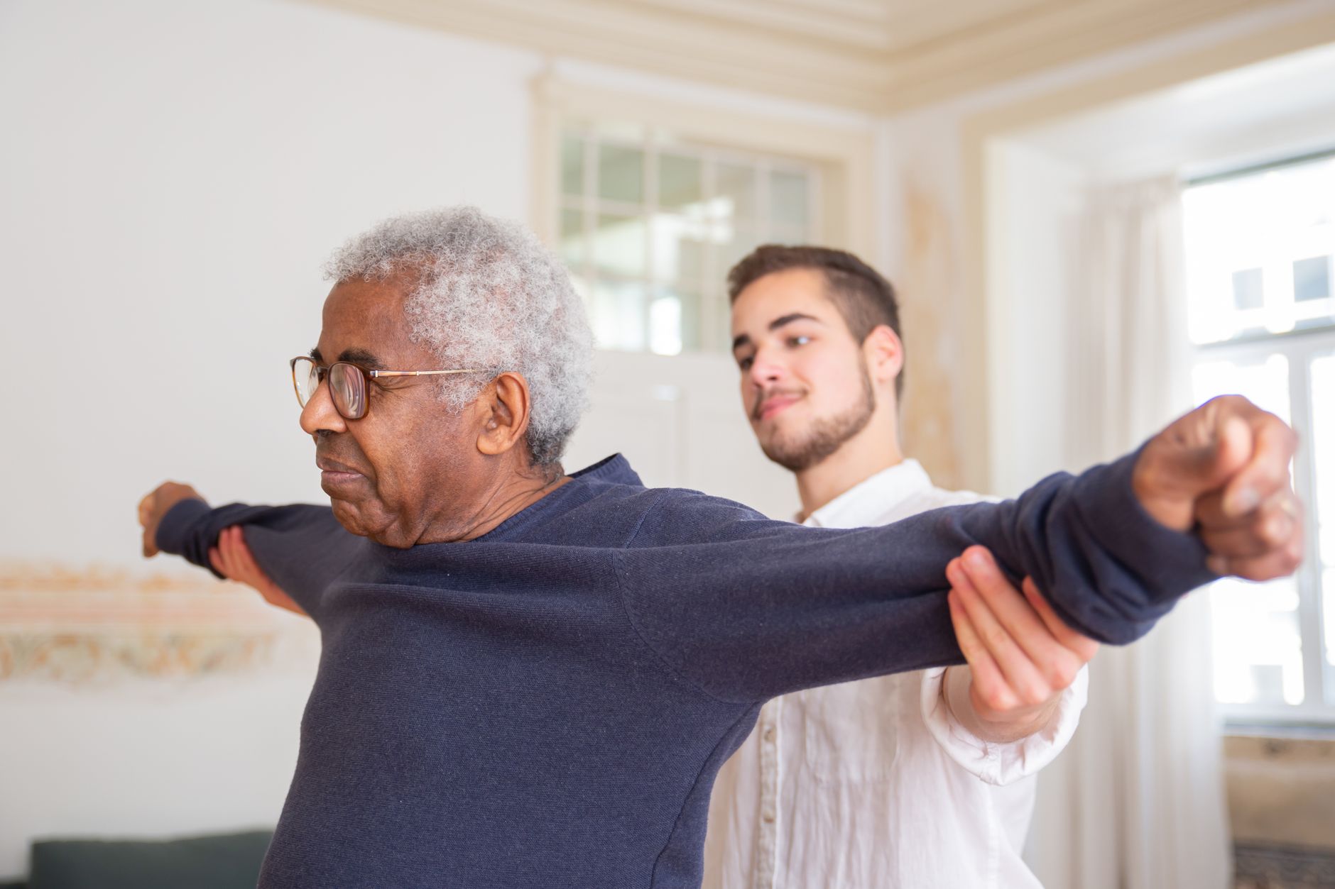A young man supports the outstretched arms of an older man in glasses during a stretching exercise in a bright room