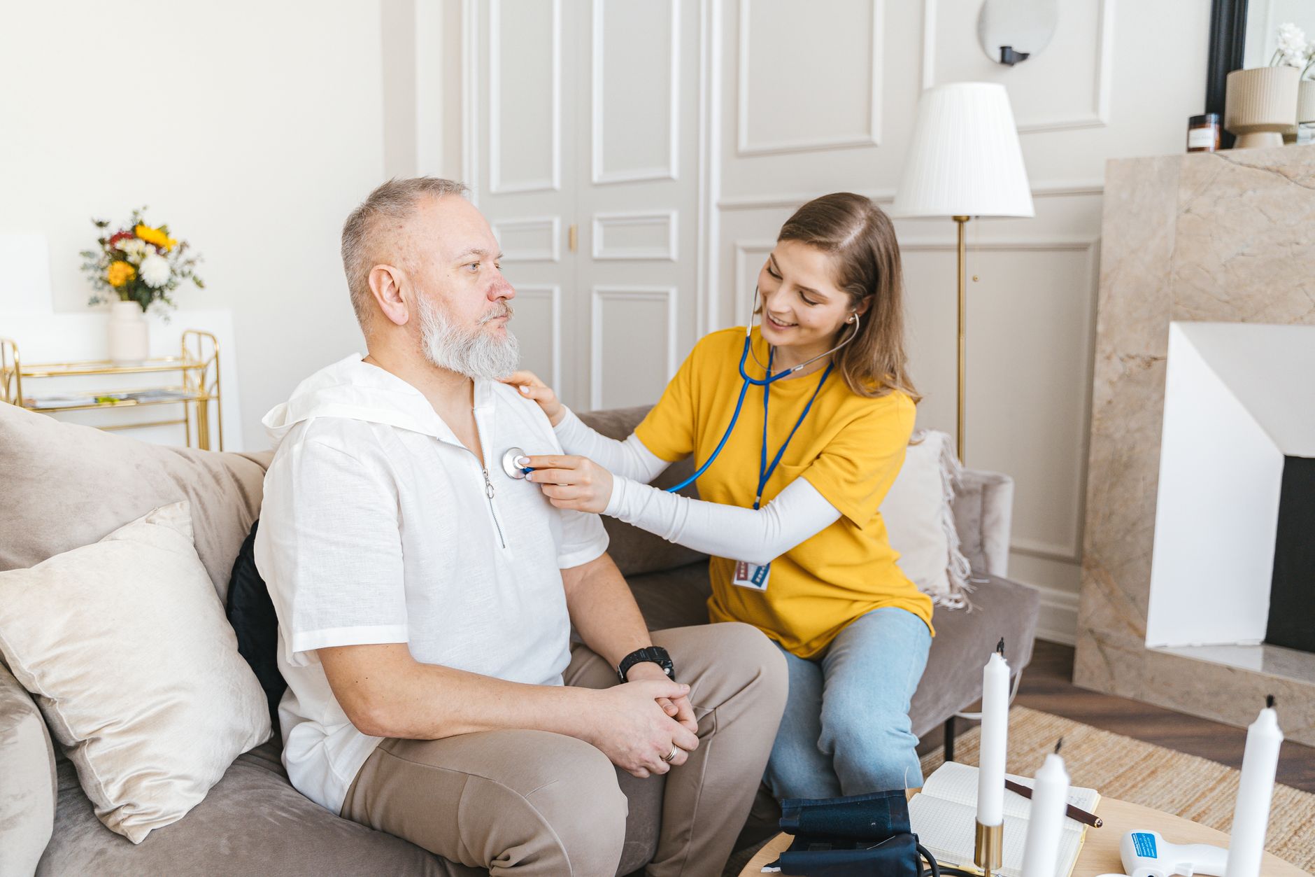 A smiling nurse in a yellow shirt listens to an older man's chest with a stethoscope as he sits on his living room sofa