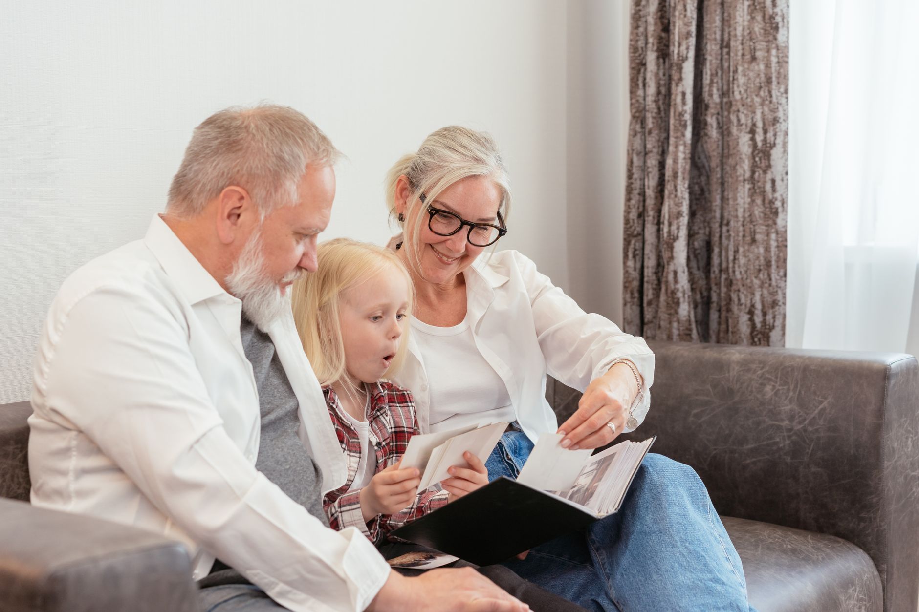 An older man and woman sit on a sofa looking through a photo album with their young granddaughter