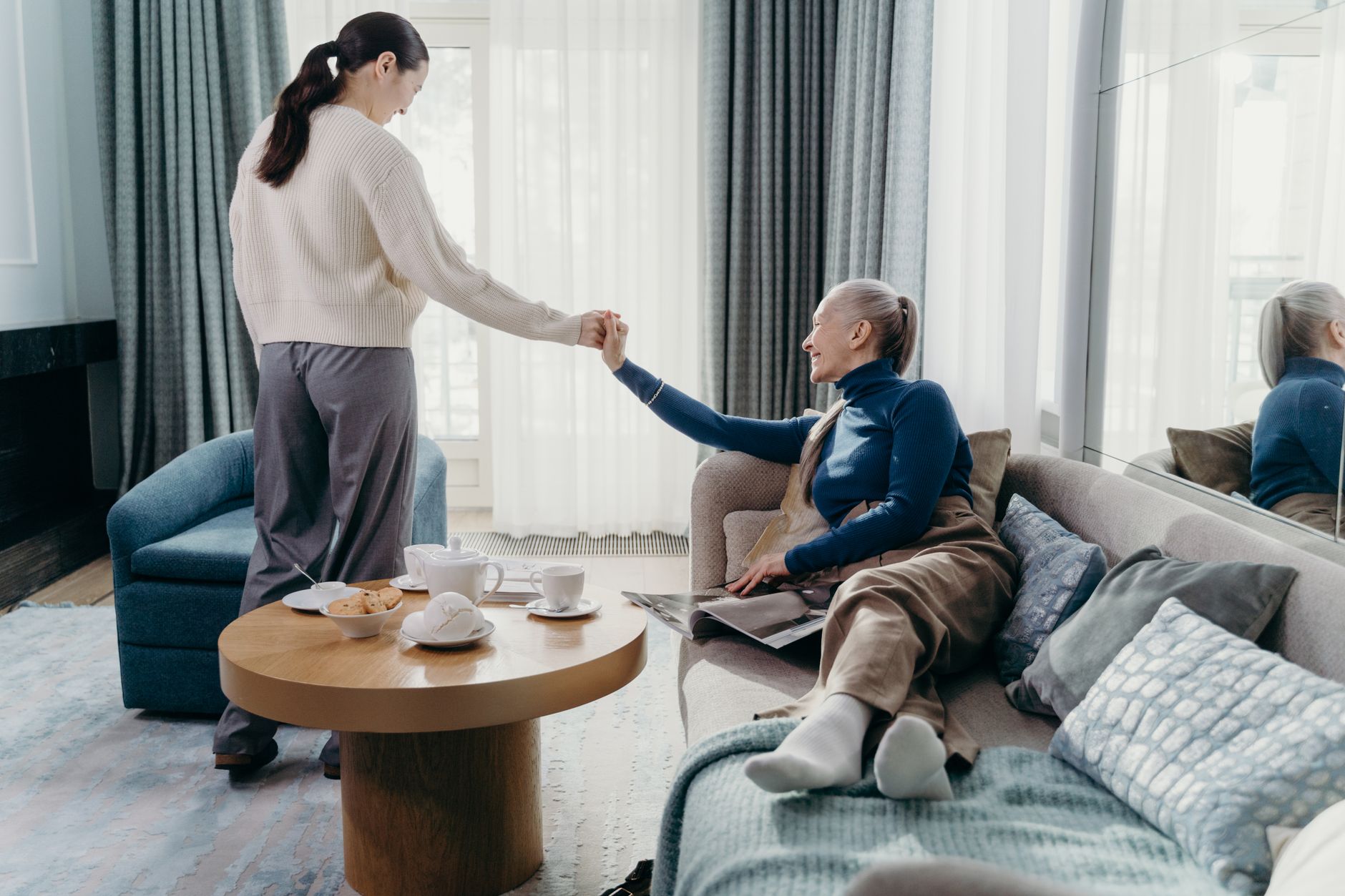 An adult daughter holds her smiling mother's hand as the mother relaxes on a sofa beside a table set for tea