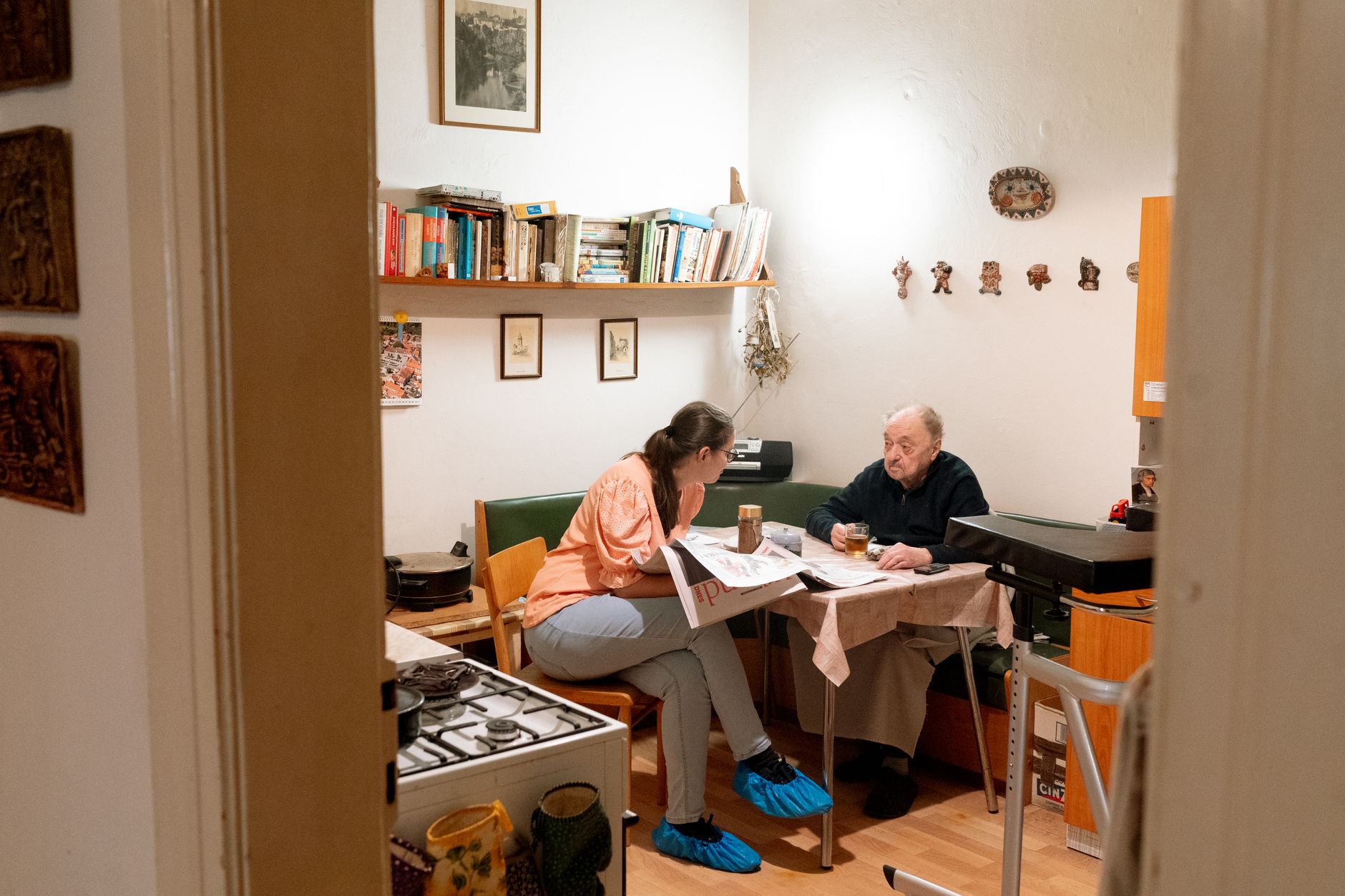 A young woman sits at a kitchen table with an older man, talking over cups of tea, with a walker beside the table