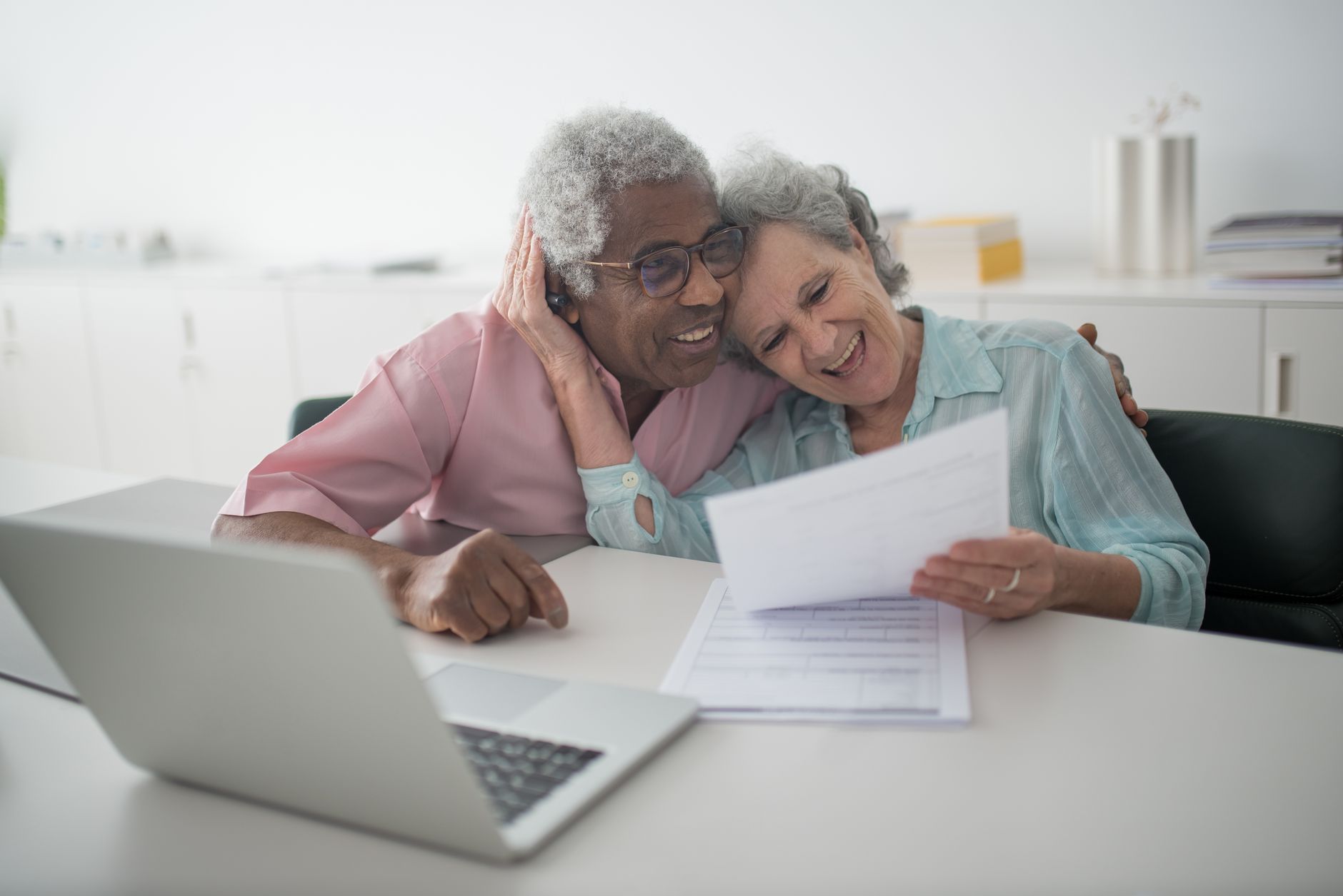 An older couple sit close together at a desk, smiling as they look over papers beside a laptop
