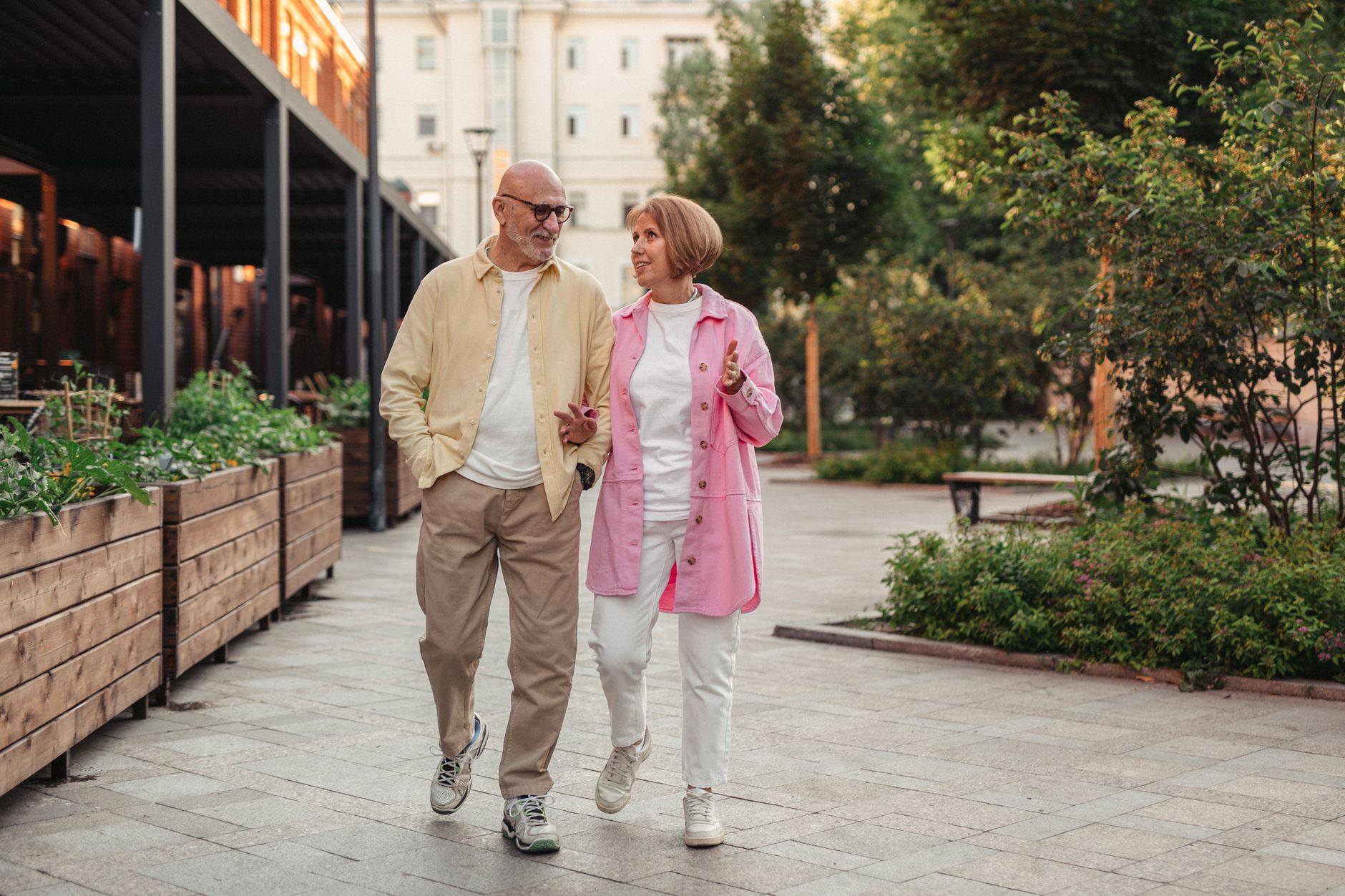 An older man in a yellow shirt and an older woman in a pink jacket walk and talk along a tree-lined city path