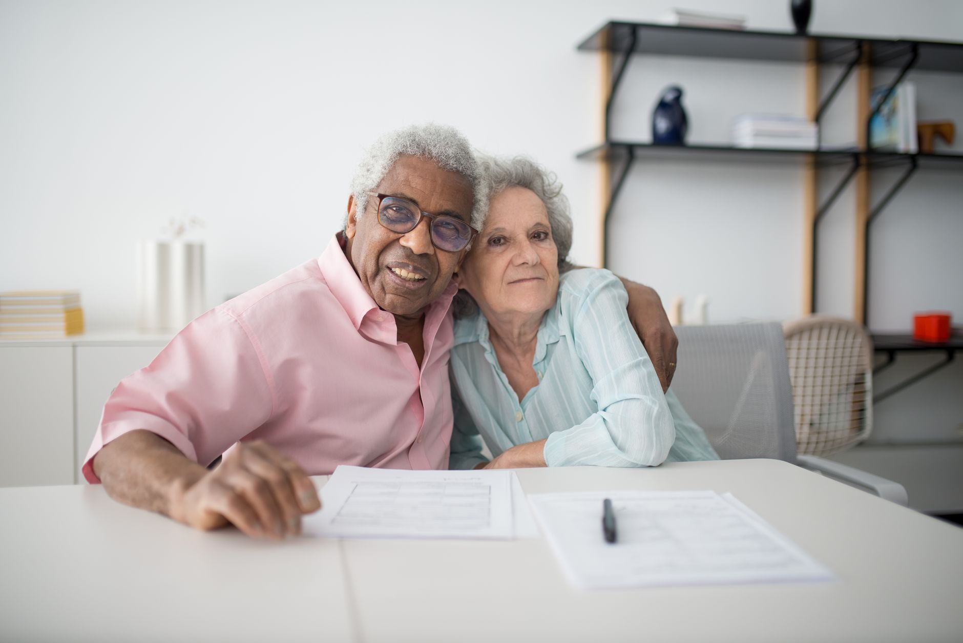 An older man in a pink shirt and an older woman sit close together at a white table with papers and a pen in front of them