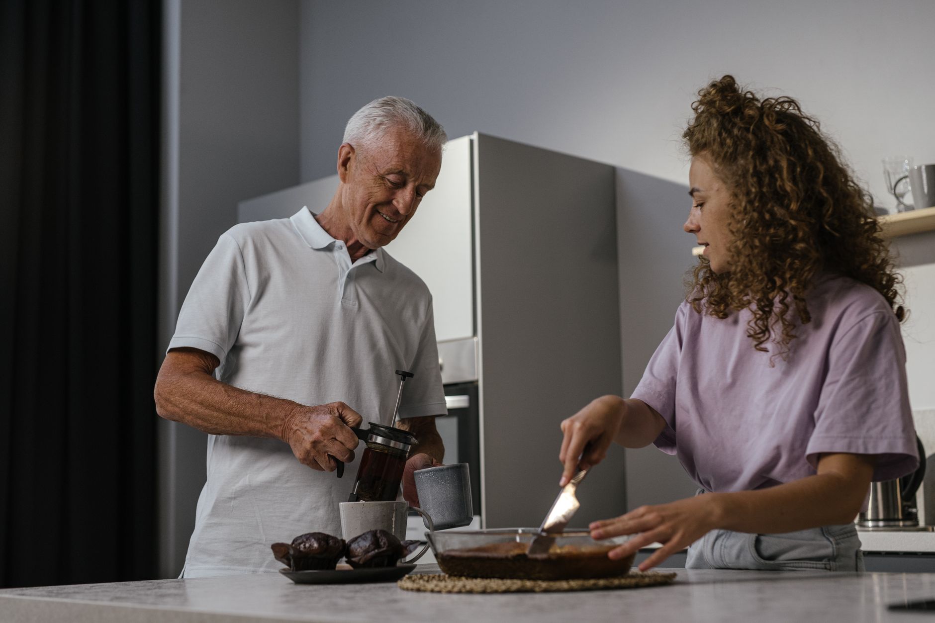 An older man pours tea from a press into a mug while a young woman cuts a homemade cake at a kitchen counter