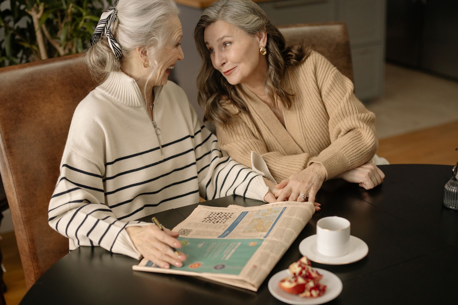 Two older women in knit sweaters laugh together at a dark wooden table with a newspaper, a white coffee cup and a plate of pomegranate
