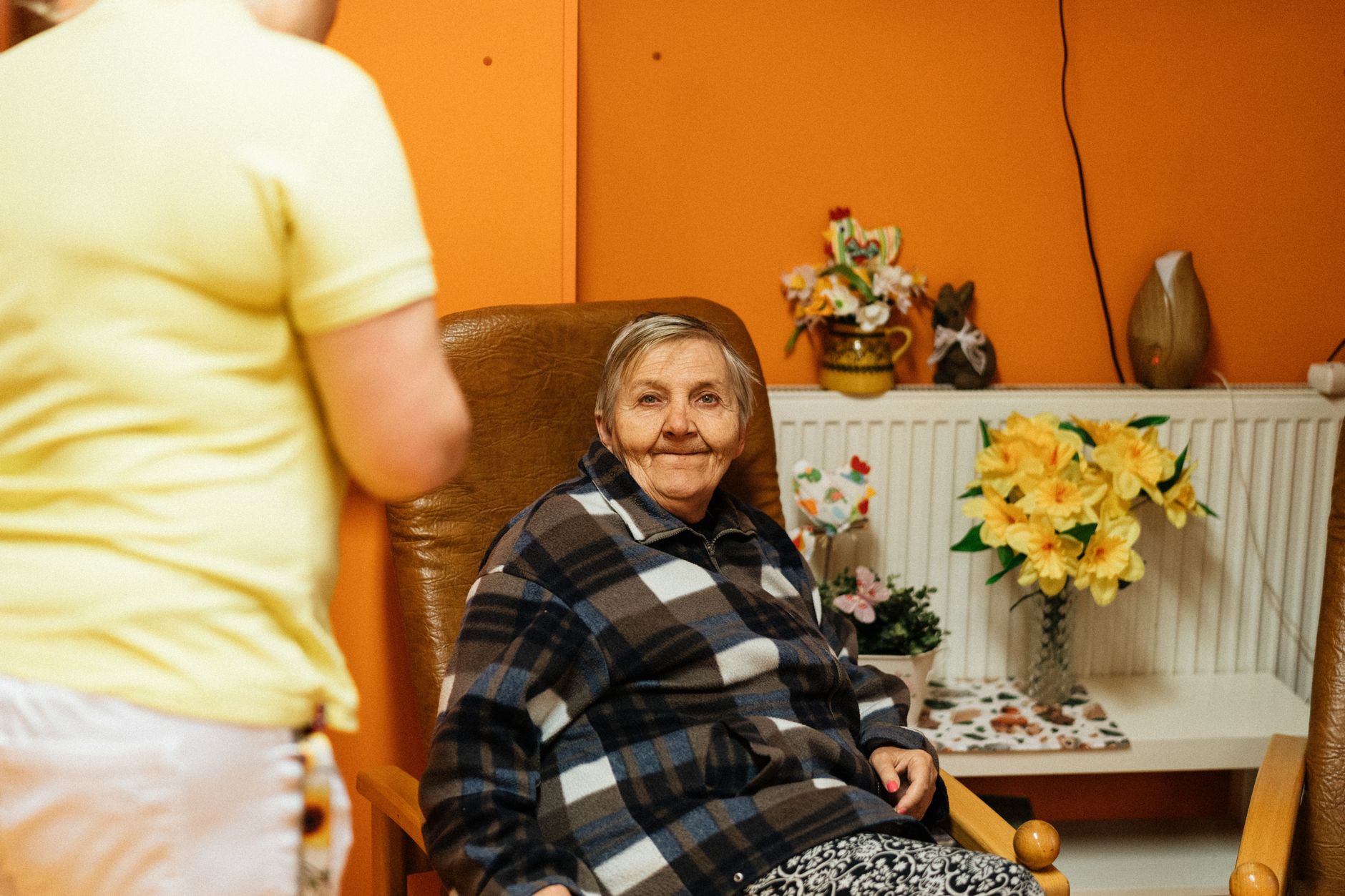 A smiling older woman in a plaid fleece jacket sits in a leather armchair in a bright orange room with flowers, while a caregiver in a yellow shirt stands nearby