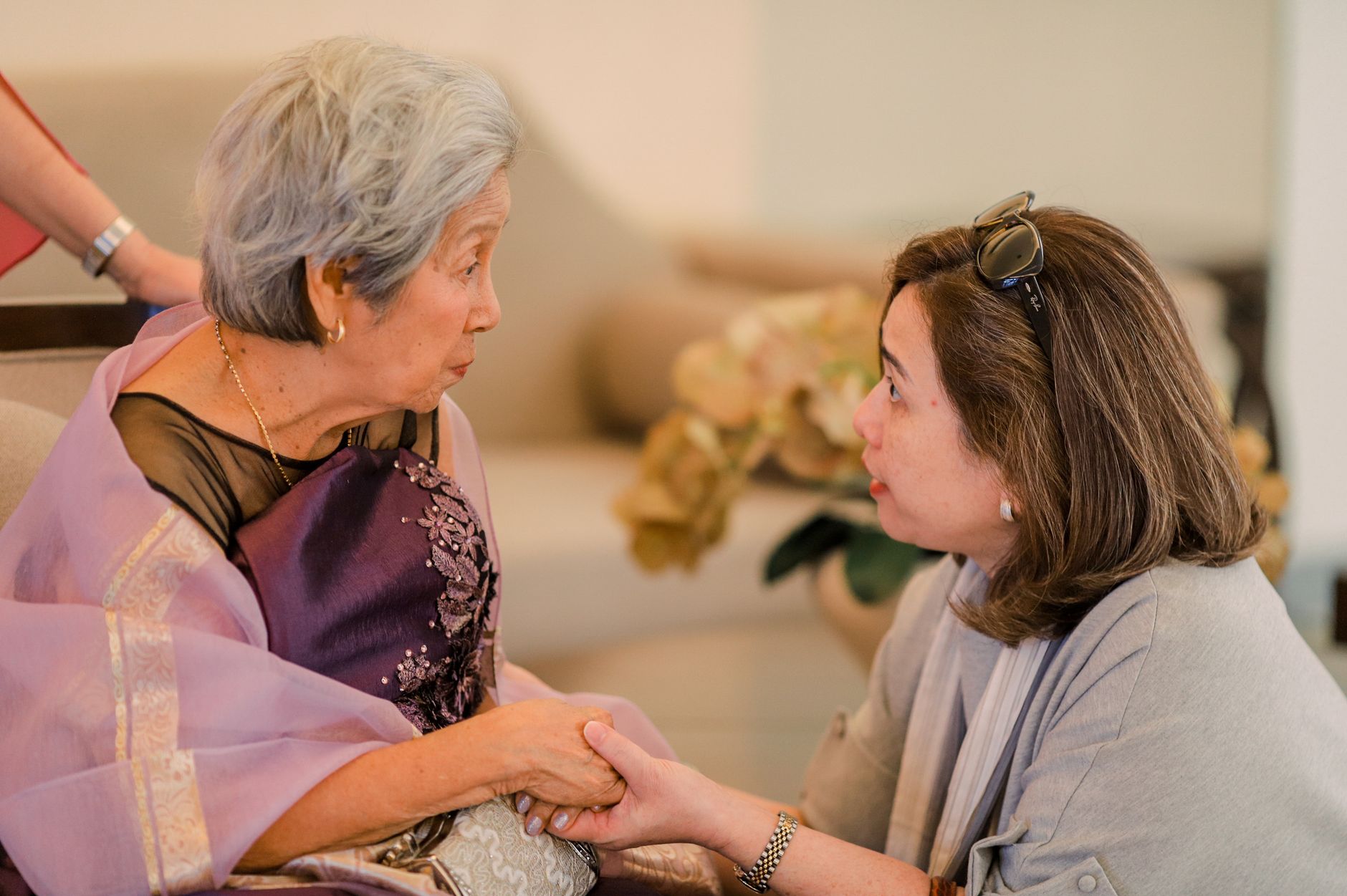 An adult daughter kneels beside her mother, holding her hands as they talk face to face