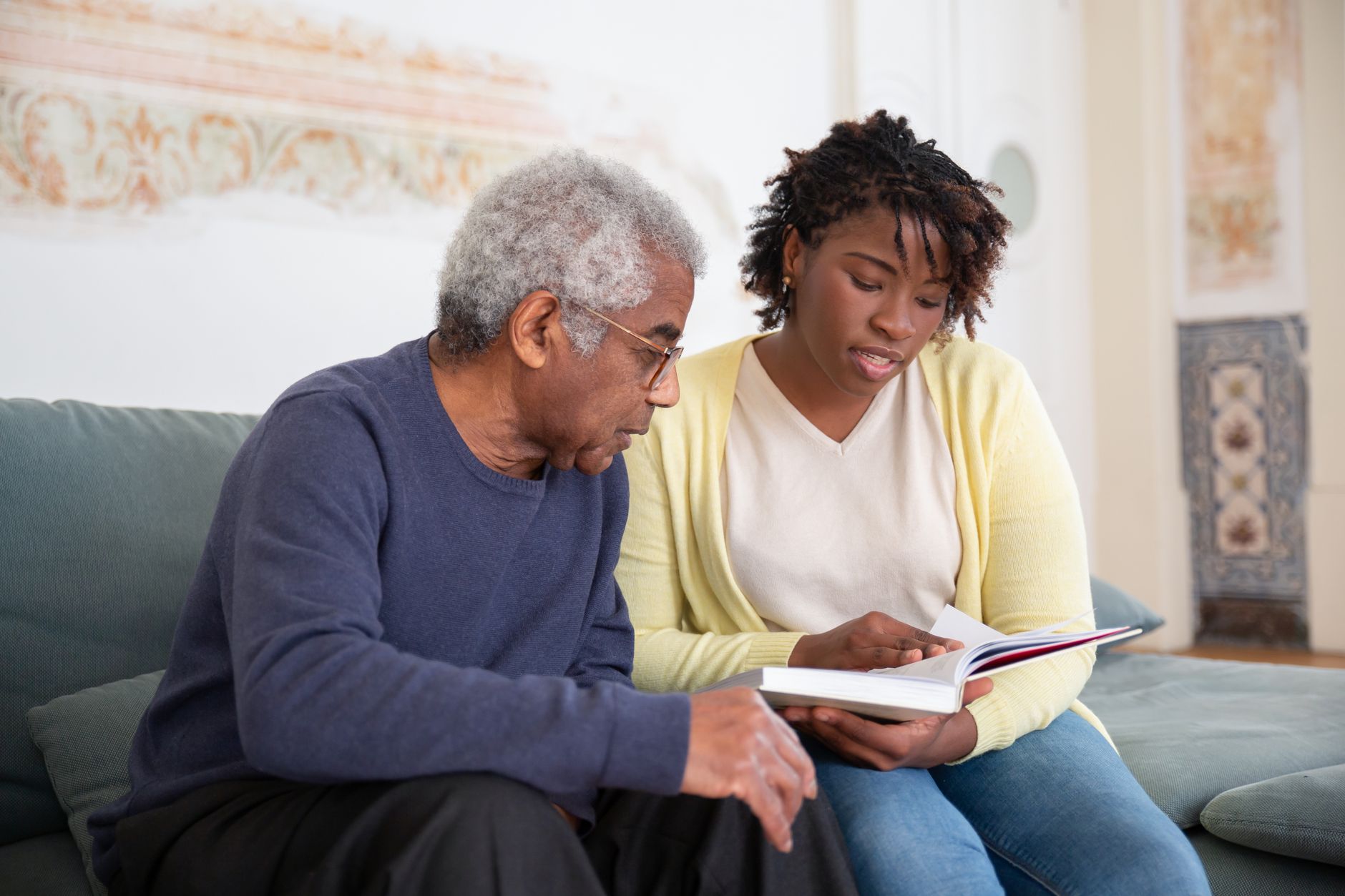 An older man and a younger woman sit side by side on a sofa, reading through a book together