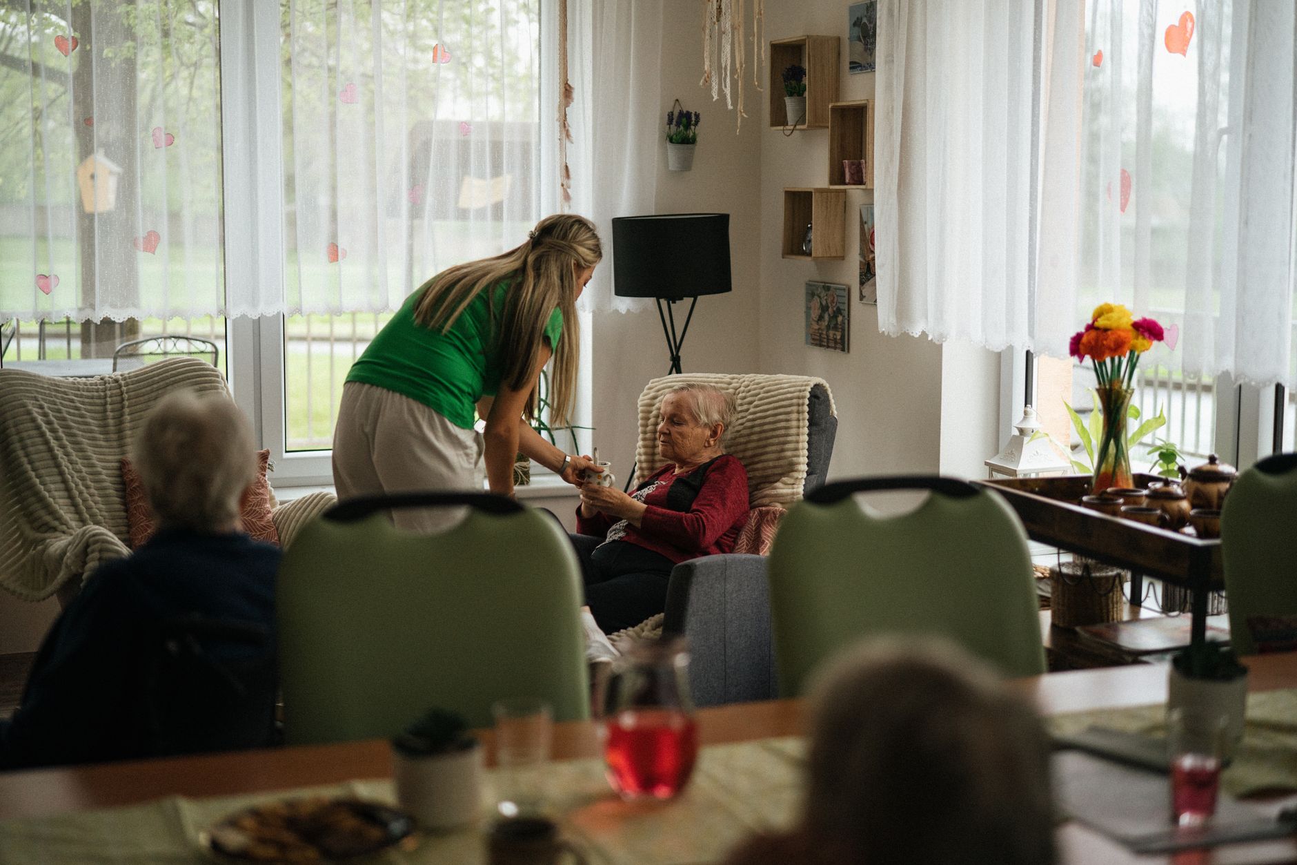 A caregiver leans down to hand a mug to an older woman resting in an armchair in a bright day room