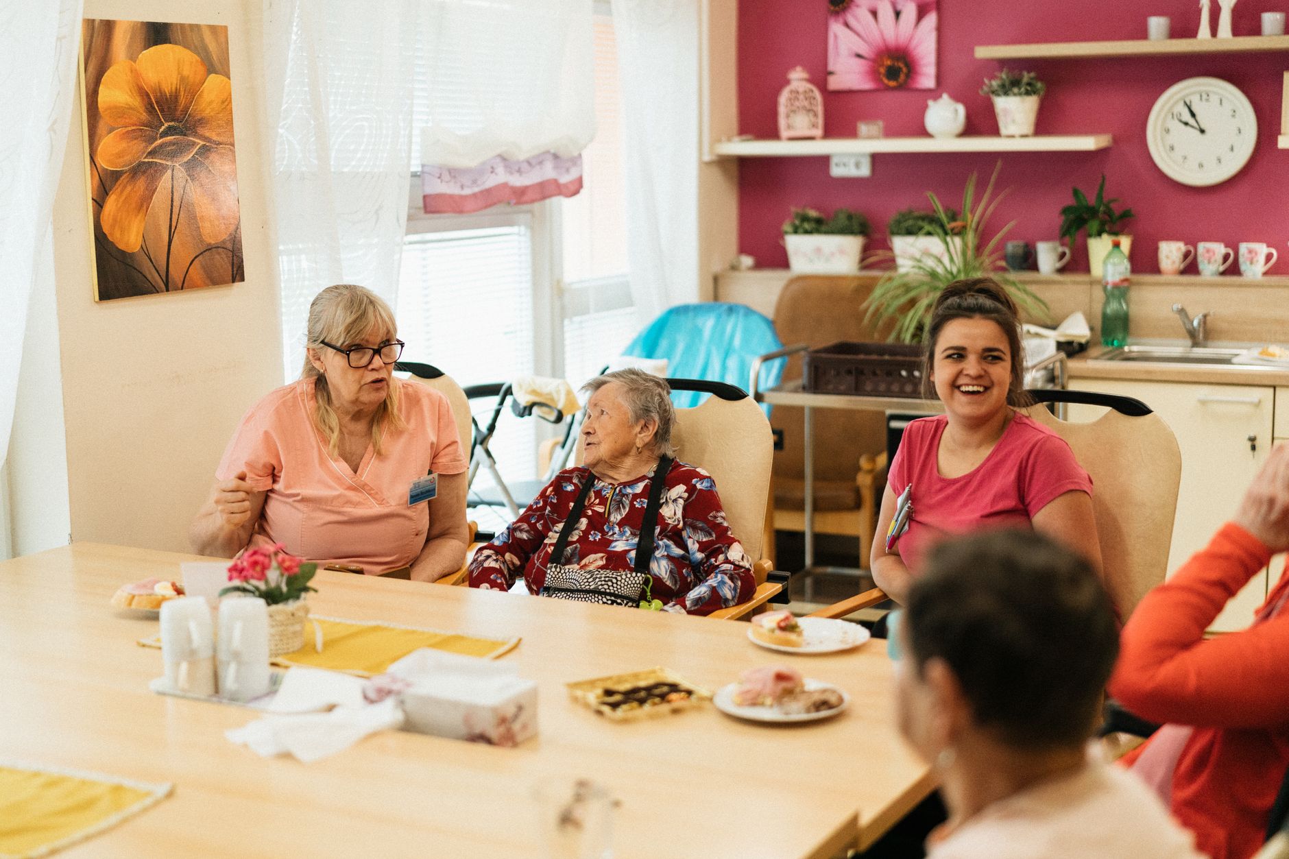 A nursing assistant chats with an older woman at a dining table while another staff member laughs nearby