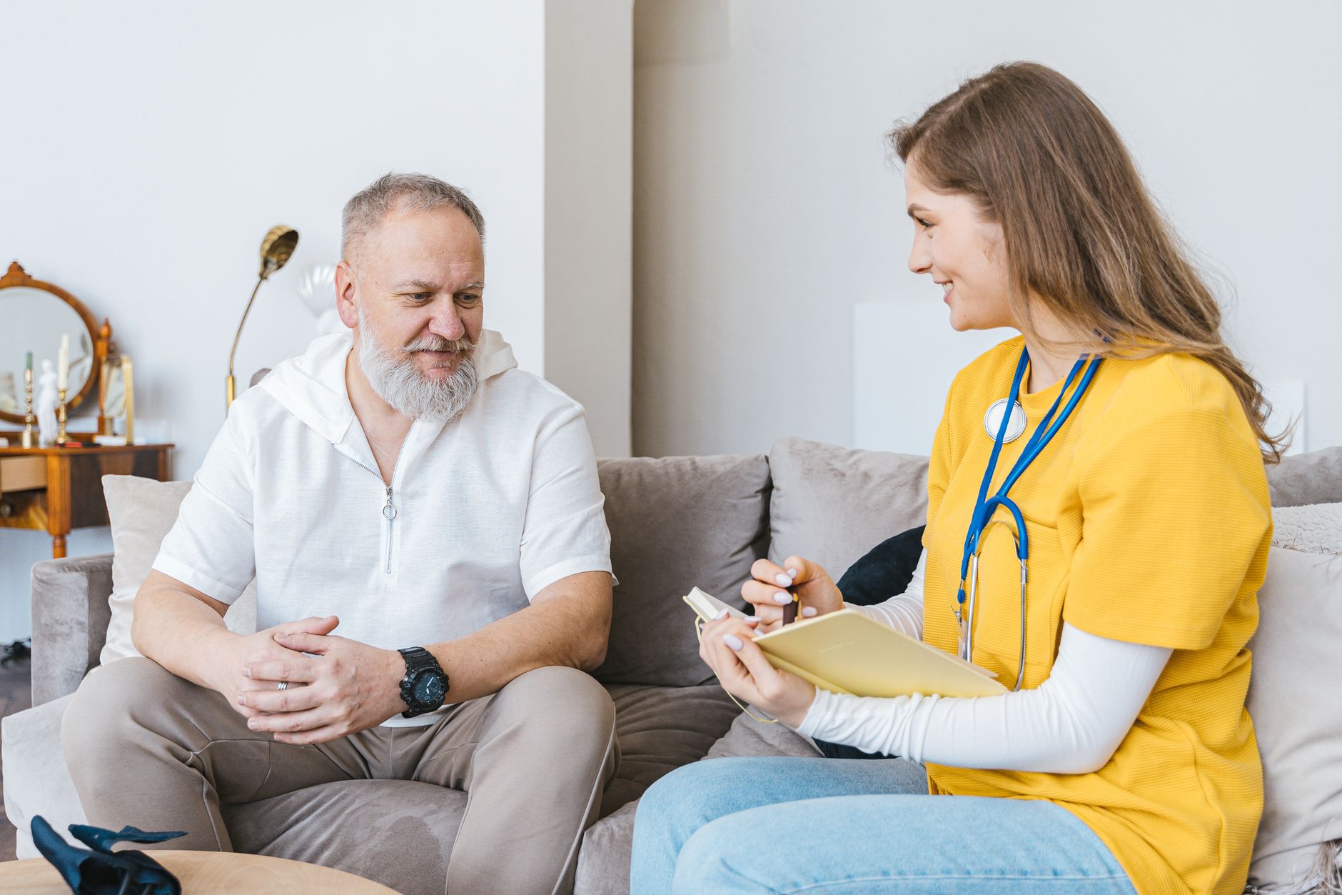 A nurse in a yellow top sits on a sofa with an older man, taking notes as they talk