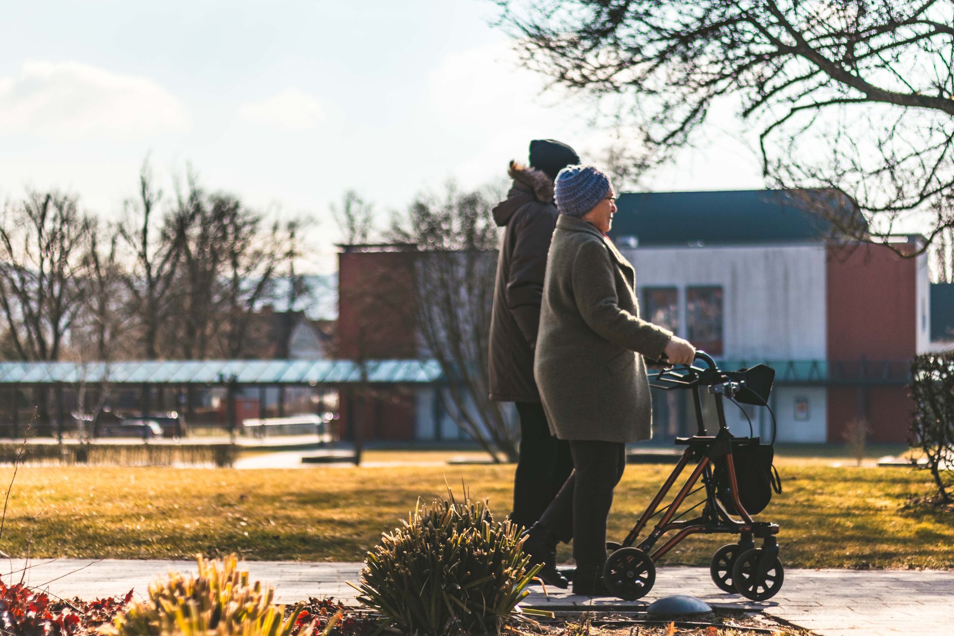 An older woman in a knit hat walks with a rollator beside a companion along a sunny park path