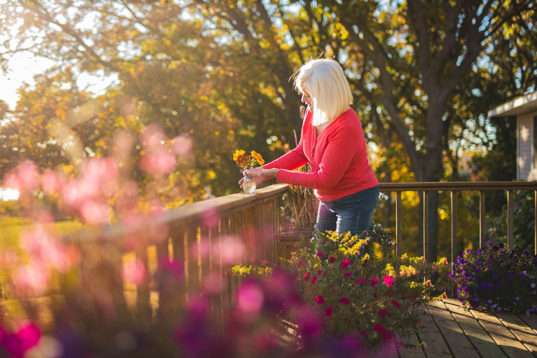 A white-haired woman in a red sweater arranges a small vase of flowers on a sunny deck surrounded by potted plants