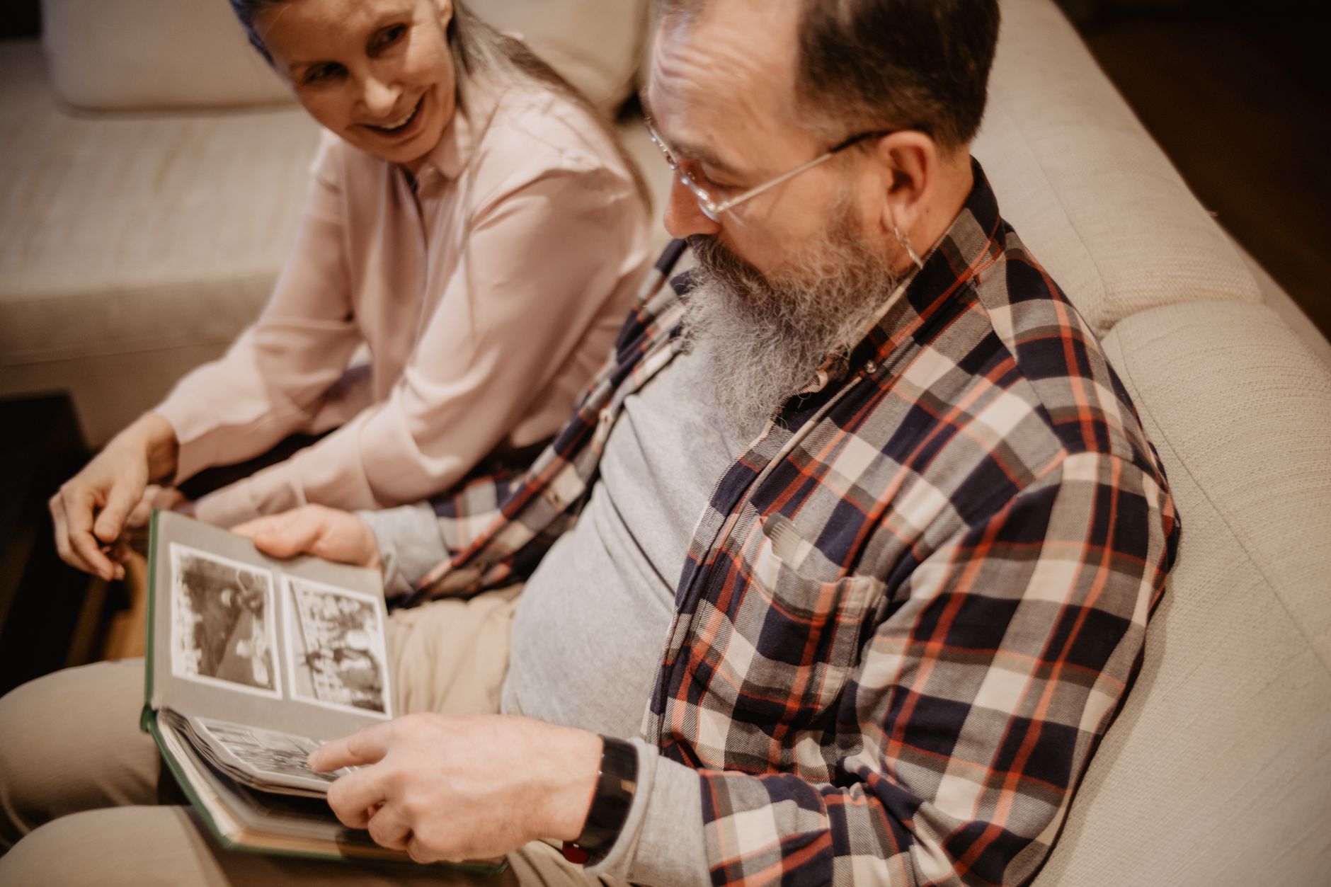 An older bearded man in a plaid shirt looks through a photo album on a sofa while an older woman beside him smiles